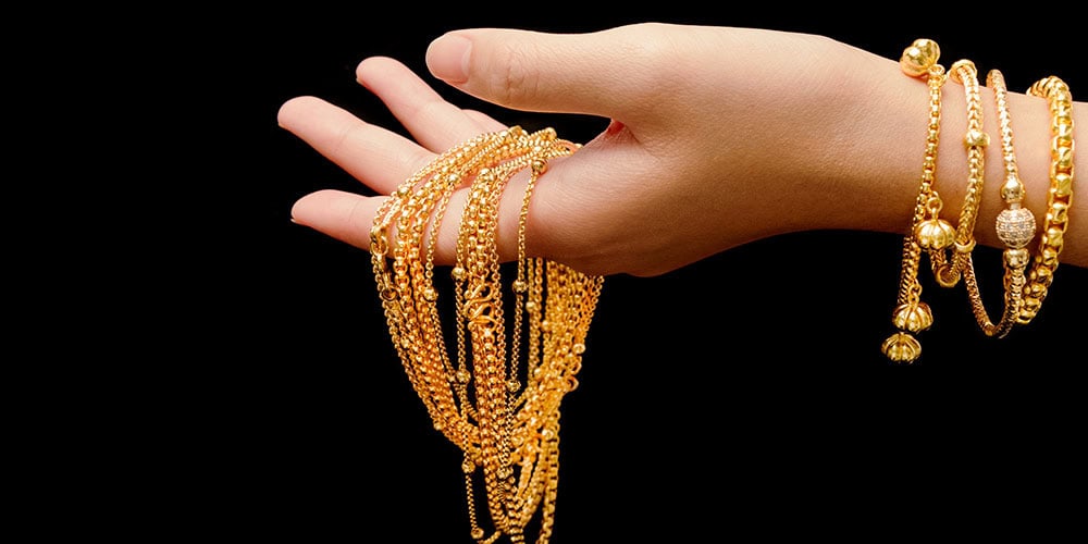 Shiny gold jewelry collection featuring layered necklaces and bracelets with intricate designs, displayed on a hand against a black background.