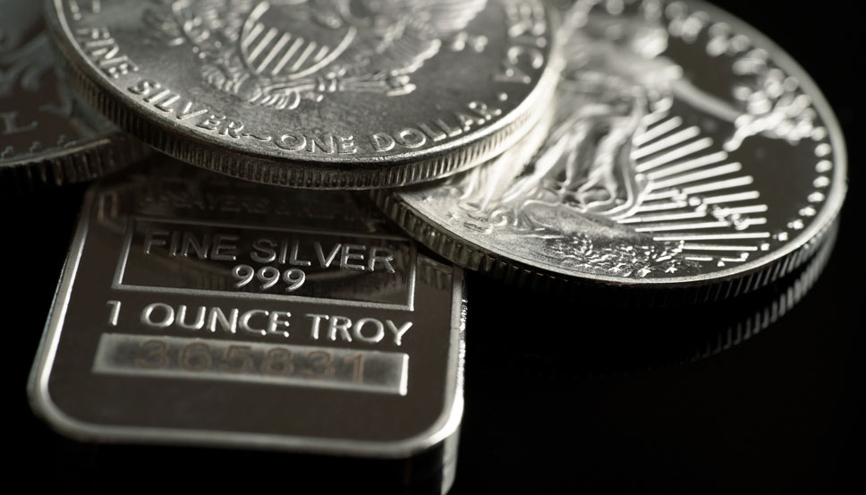 A professional jeweler is carefully examining various silver items, including coins and bars, using precision tools to assess their quality and value in the current silver market. The jeweler's focus highlights the importance of understanding silver prices and the significance of these precious metals in investment portfolios.